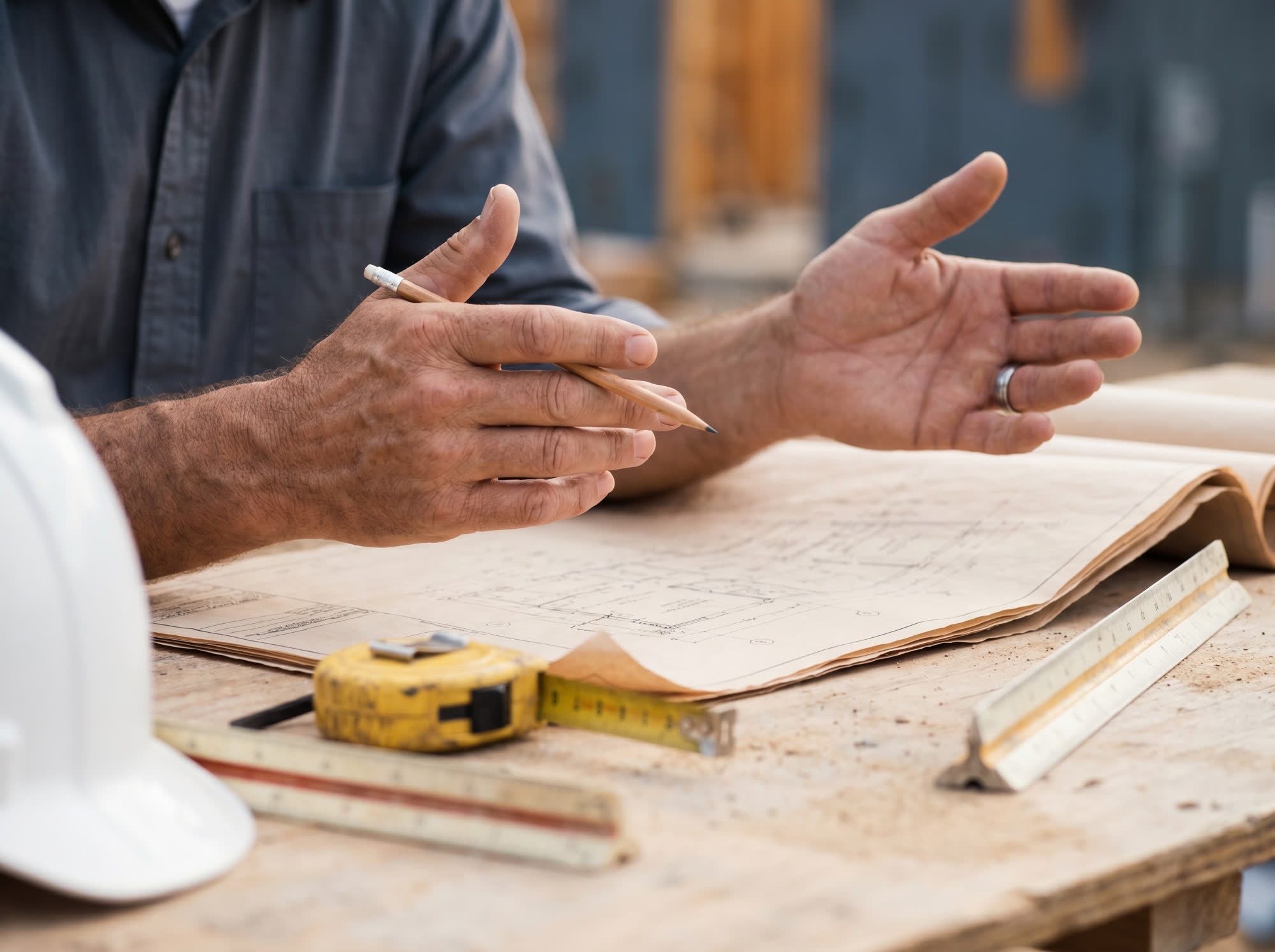 Estimator's hands reviewing rolled-out architectural blueprints on a job-site table beside a hard hat, scale ruler, and tape measure.